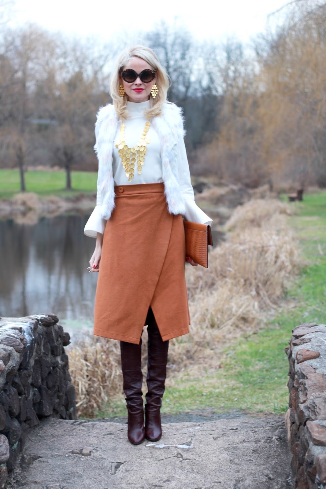 Woman in dark brown slouchy boots with a rust wrap skirt, white turtleneck and white faux fur vest