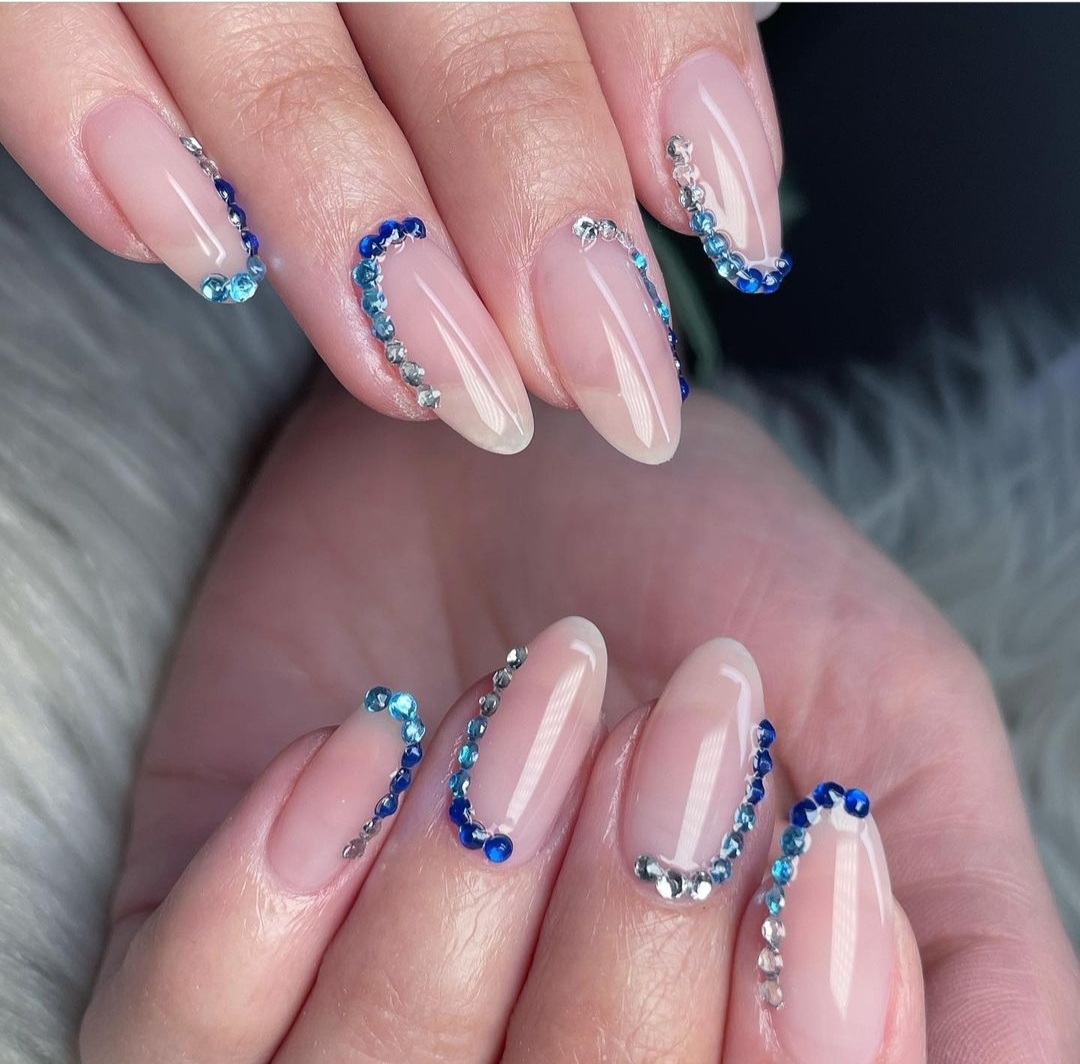 Medium almond nails in nude with French tip arcs made of blue and clear rhinestones