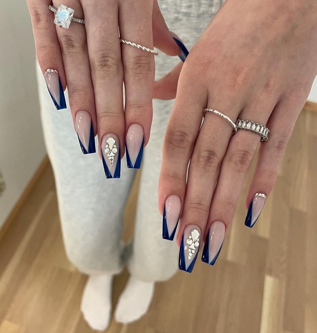 Long coffin nails with icy-blue V-shaped French tips on a nude base; rhinestone/gem clusters set at the cuticles plus one statement crystal accent nail. Real photo, no watermark. Credit page confirmed live via page-text fetch.