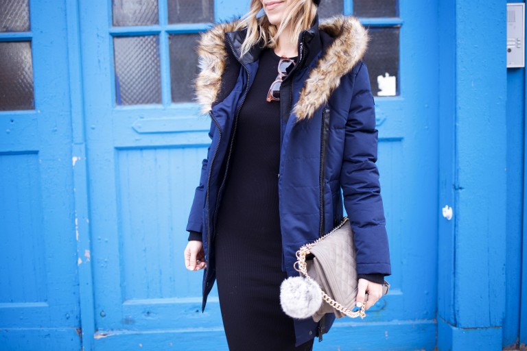 Woman in a navy fur-hood parka over a black dress with a quilted chain bag