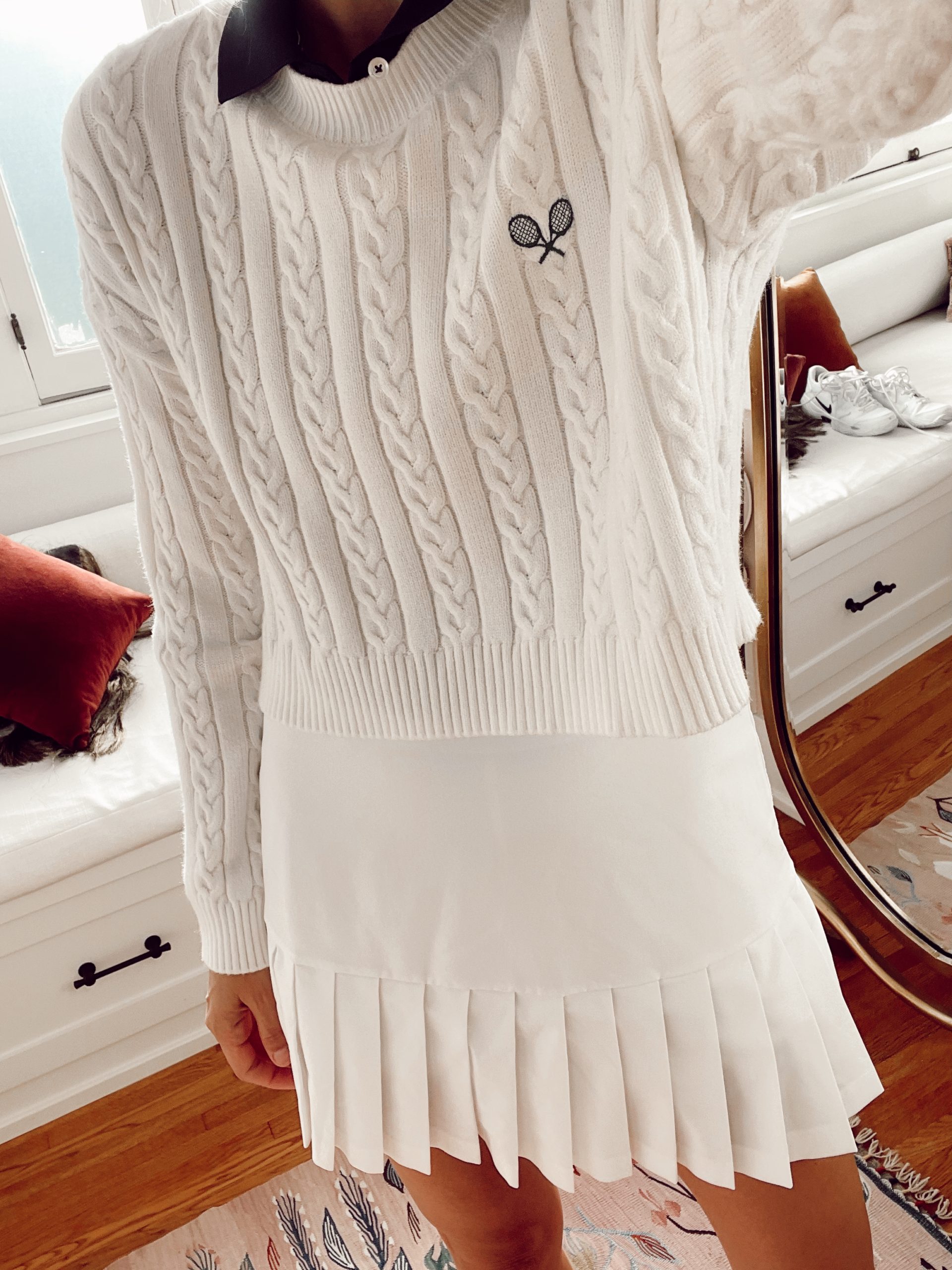 Woman wearing a white cable-knit tennis sweater with racket embroidery and a white pleated tennis skirt