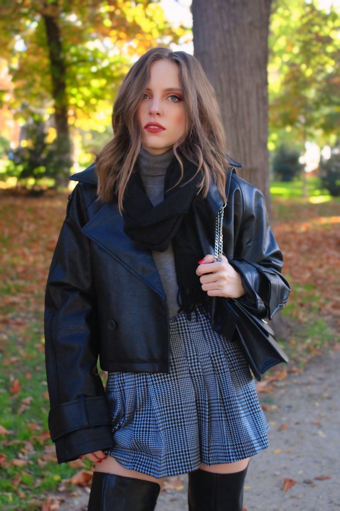 Woman wearing a black leather biker jacket with a grey turtleneck and a houndstooth pleated mini skirt with knee-high boots
