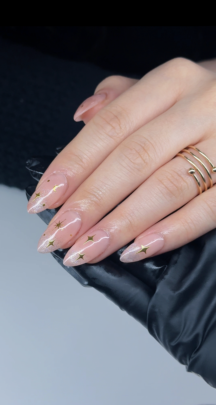 Sheer nude almond gel nails with gold star decals and a subtle glitter fade