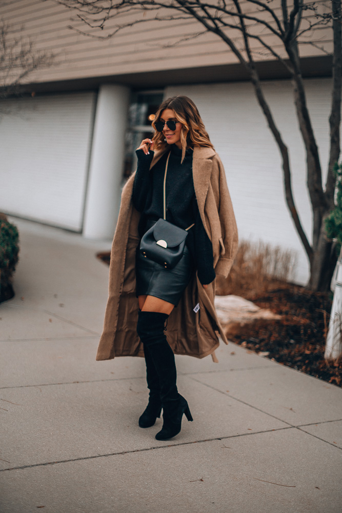 Woman in a camel coat worn open over a black sweater, black leather mini skirt and over-the-knee boots
