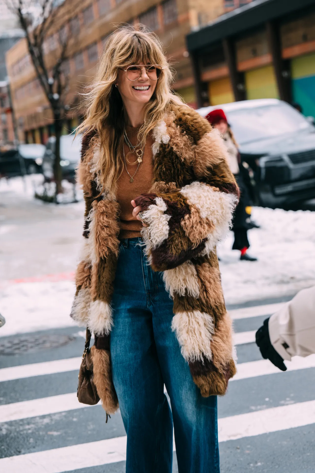 Woman wearing a brown and cream patchwork faux-fur coat with a rust knit and blue jeans in the snow