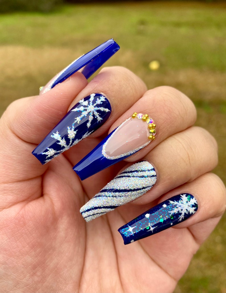 Long coffin nails in royal blue with white snowflakes, silver glitter swirls and gold rhinestone accents
