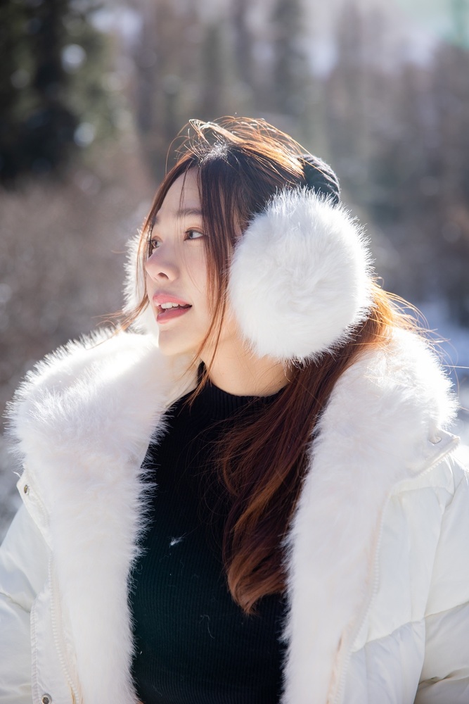 Woman wearing white faux fur earmuffs and a white puffer jacket with a fur collar in a snowy forest