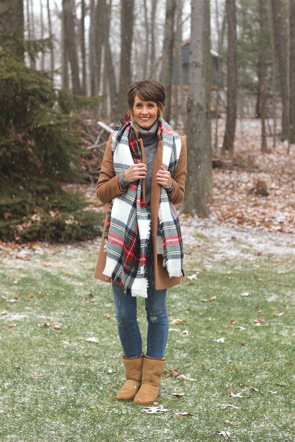 Woman in a camel coat with a red and green tartan blanket scarf, grey turtleneck and jeans in falling snow