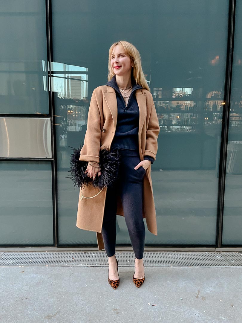 Woman wearing a camel coat over a navy jogger and sweater set with leopard-print pumps, feather bag and layered necklaces
