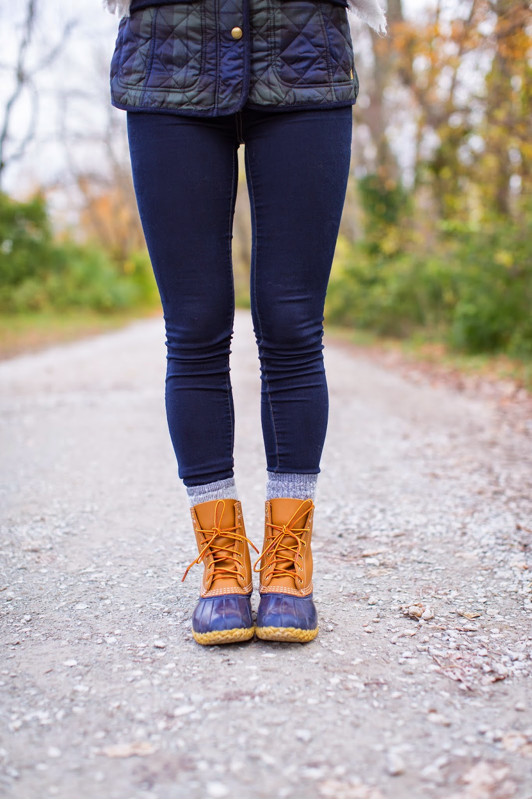 Close-up of tan and navy duck boots with dark jeans, grey socks and a quilted plaid vest