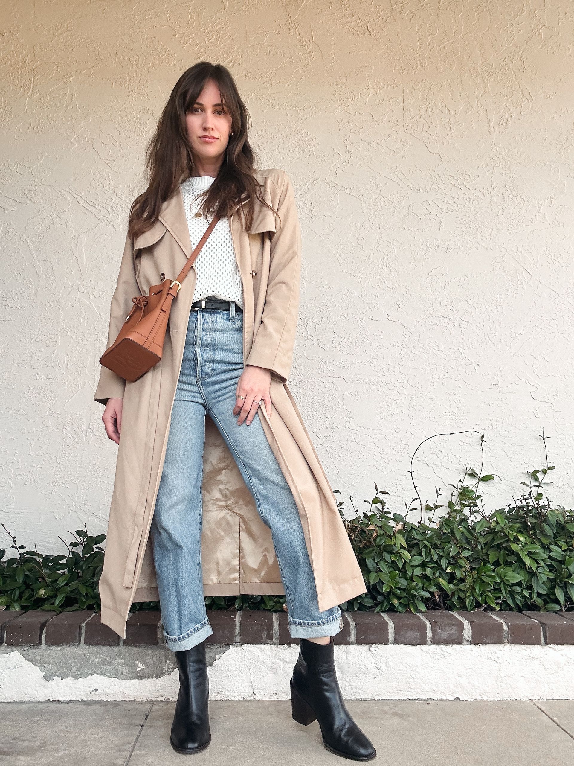 Woman in a beige trench coat with a white knit sweater, cuffed light-wash jeans and black ankle boots
