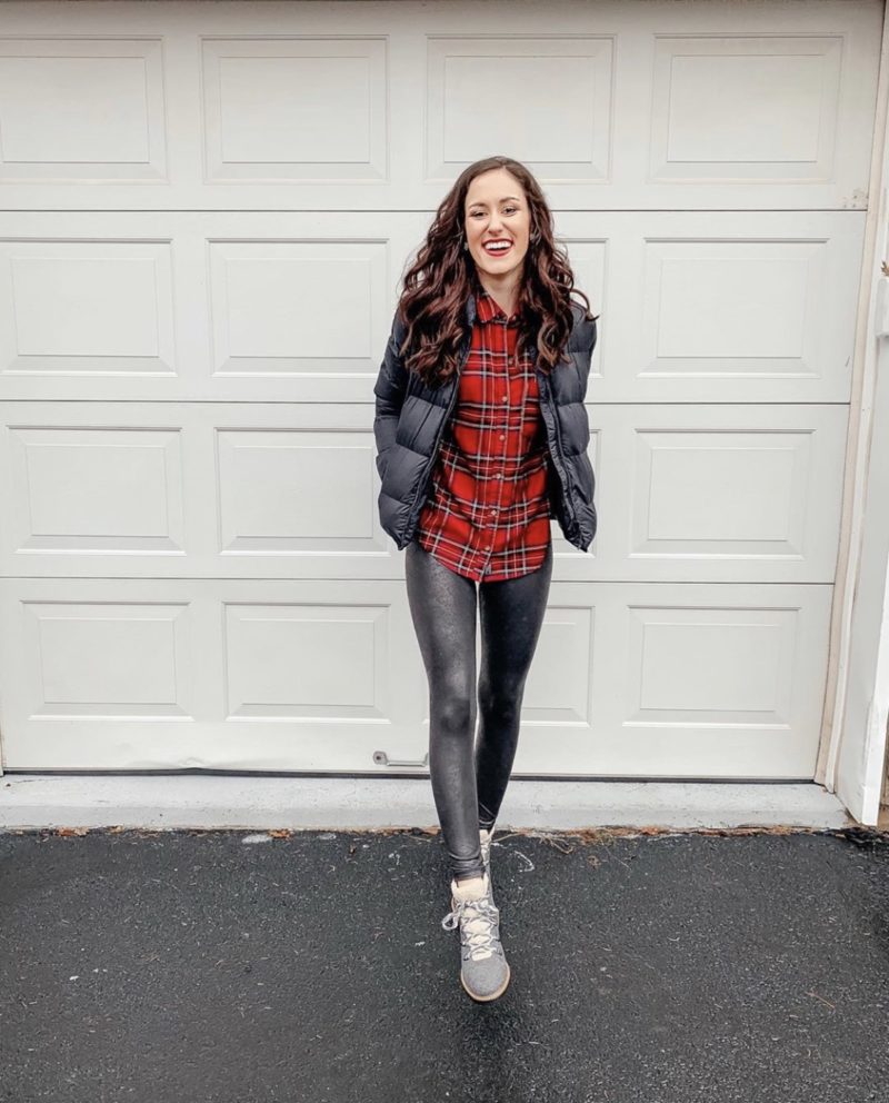 Woman in a red plaid flannel shirt, black puffer vest, dark coated leggings and grey lace-up boots