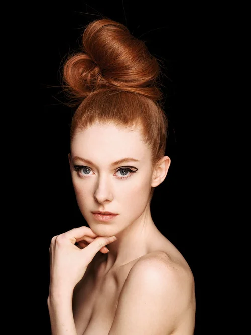 Front studio portrait of a high, tousled messy bun on vivid copper-red hair against a black backdrop.