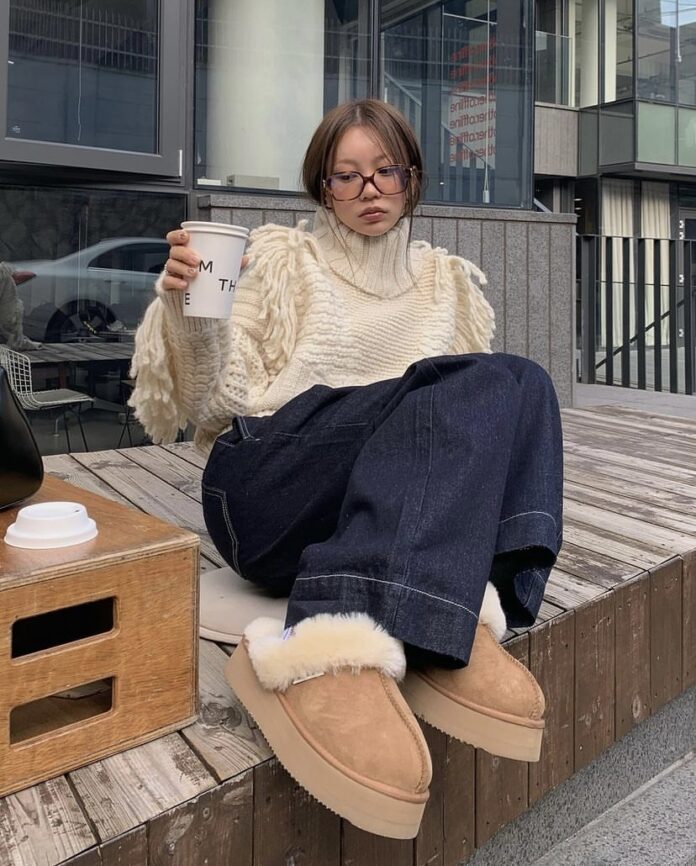 Woman in a chunky cream turtleneck and wide-leg jeans wearing chestnut platform ugg slippers