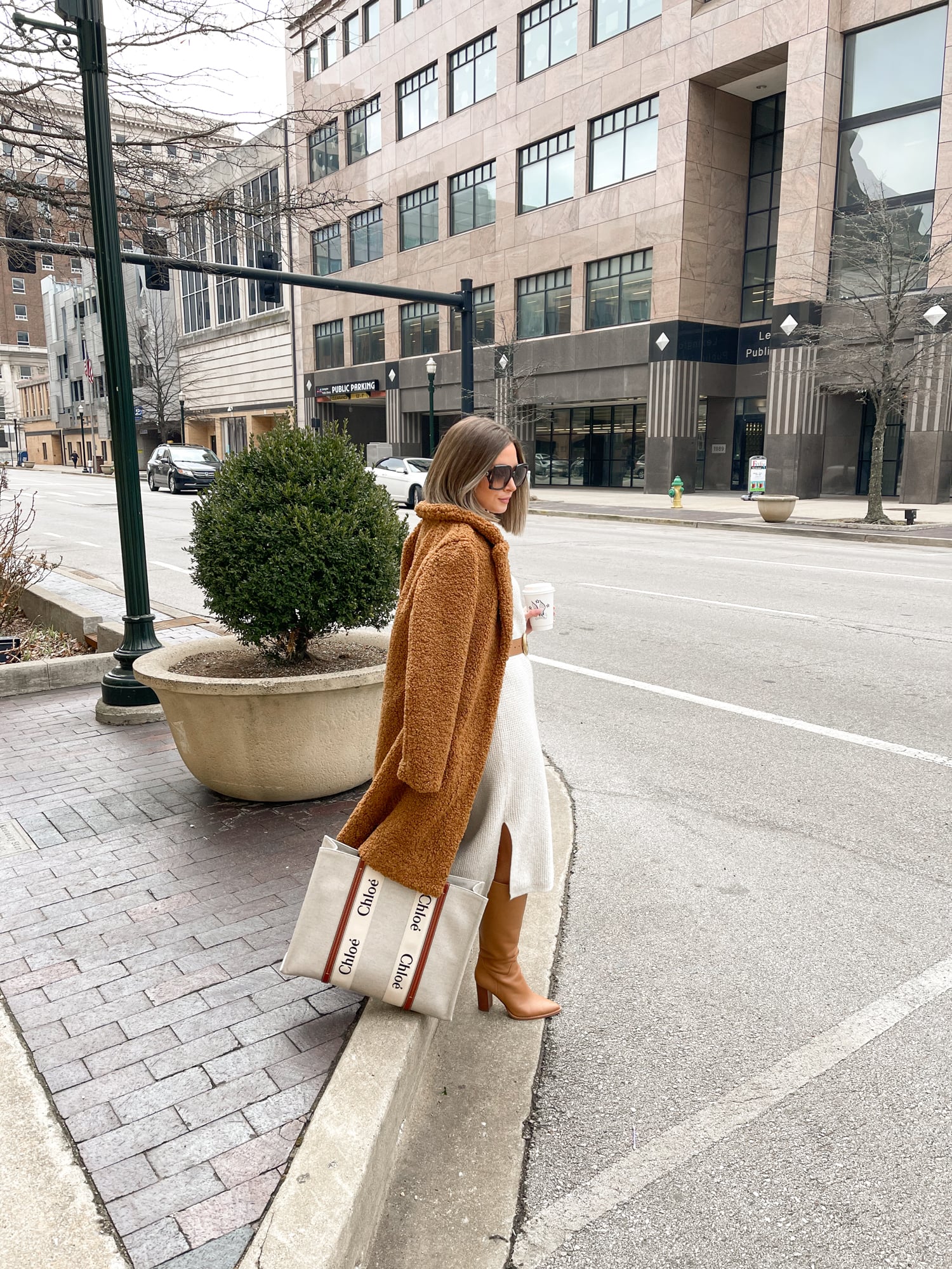 Woman in a camel teddy coat with a white knit midi dress, tan knee-high boots and a tote bag