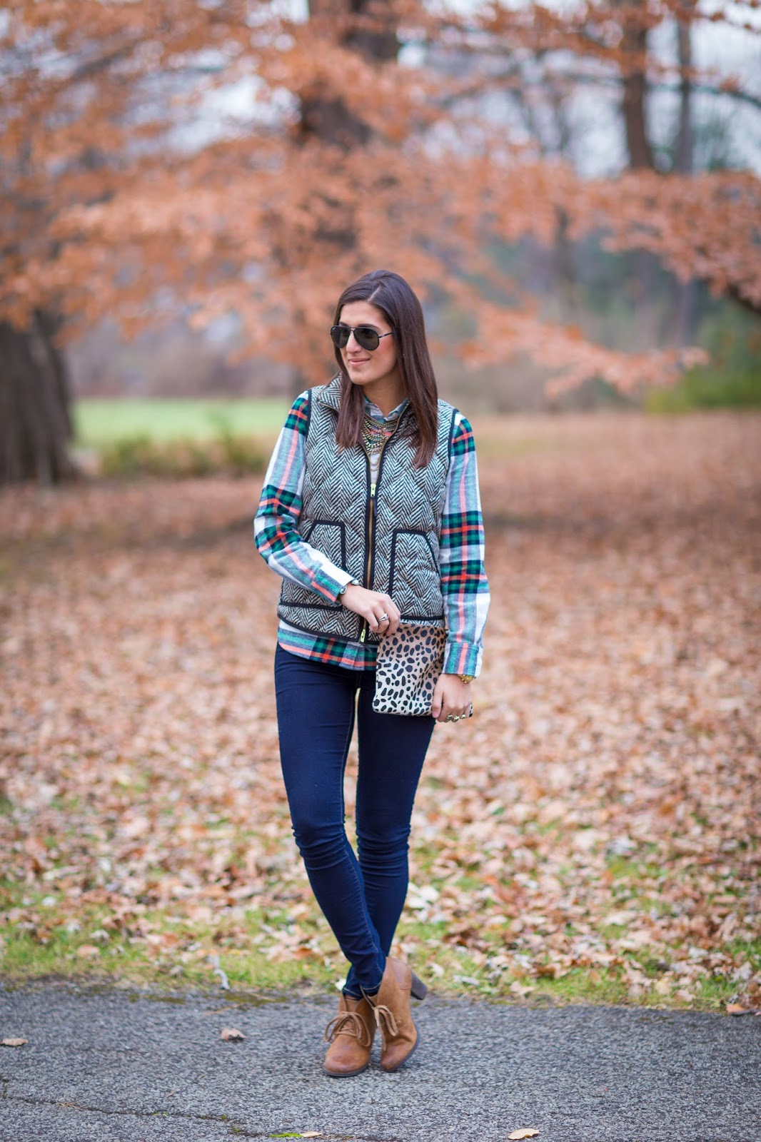 Woman wearing a herringbone quilted vest over a green and red plaid flannel shirt with dark jeans and tan booties in a park