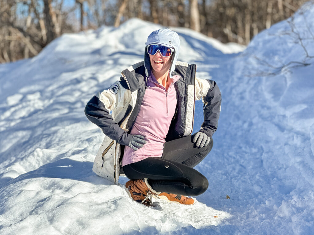Woman in a pink quarter-zip base layer sitting in deep snow on a hike