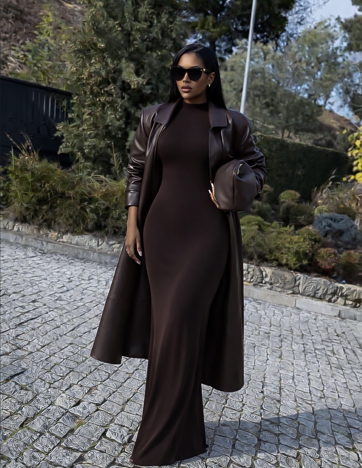 Woman in a chocolate brown bodycon maxi dress with a brown leather trench coat, sunglasses and a clutch