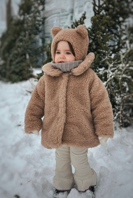 A toddler wearing a caramel teddy bear-ear coat with cream ribbed leggings standing in the snow