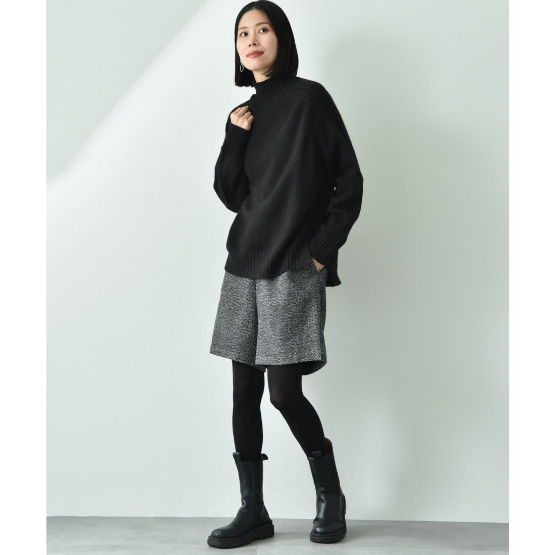 Woman wearing a black turtleneck sweater, grey tweed shorts, black tights and black chunky ankle boots