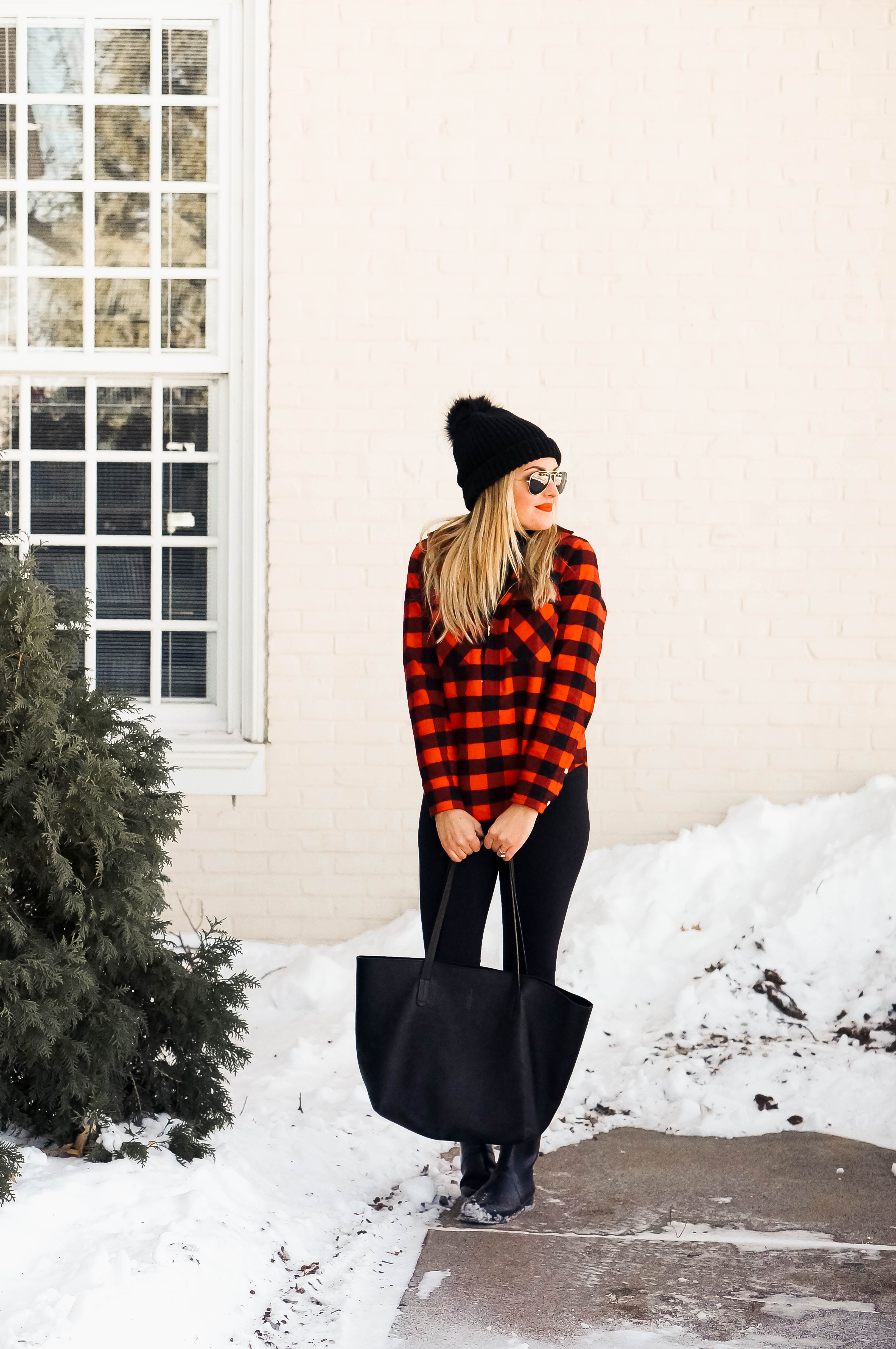 Woman in a red buffalo plaid shirt with black leggings, black boots, and a black beanie in the snow