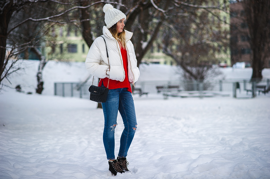Woman in a white puffer, red knit top and jeans with black lace-up winter boots in a snowy park