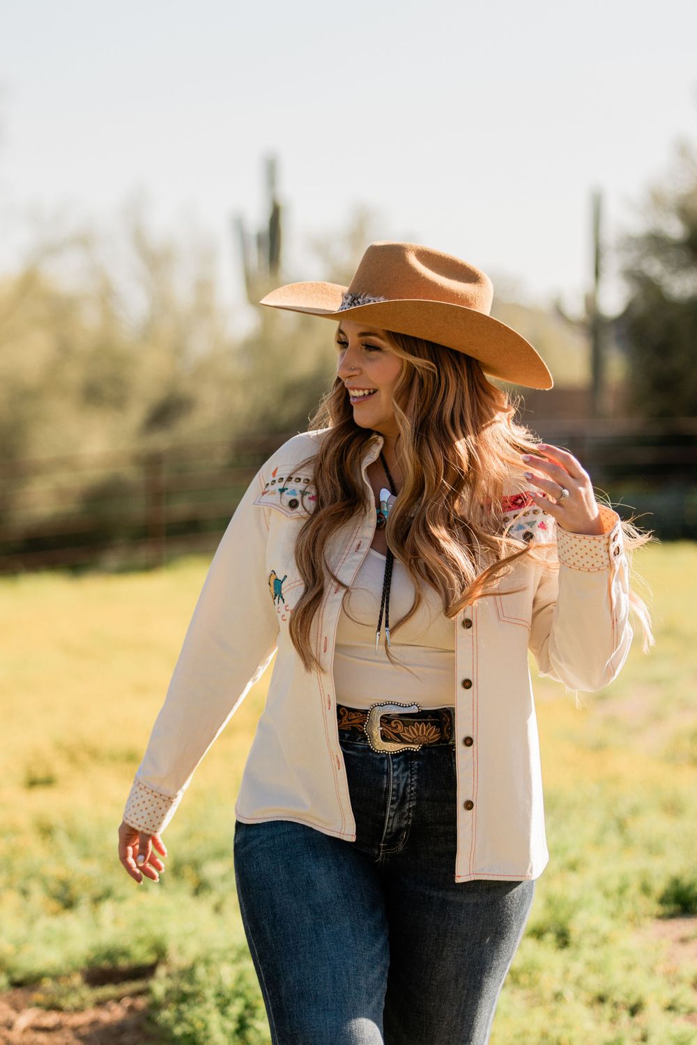 Woman in a cream embroidered western shirt with a tooled belt and tan cowboy hat in a desert field