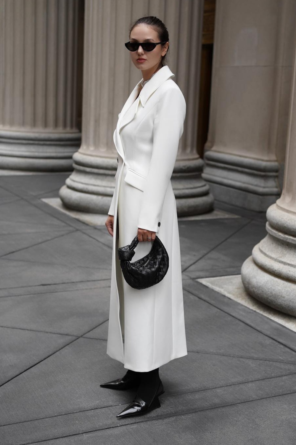 A crisp white long coat worn open over dark layers, finished with pointed black boots and a woven black handbag.