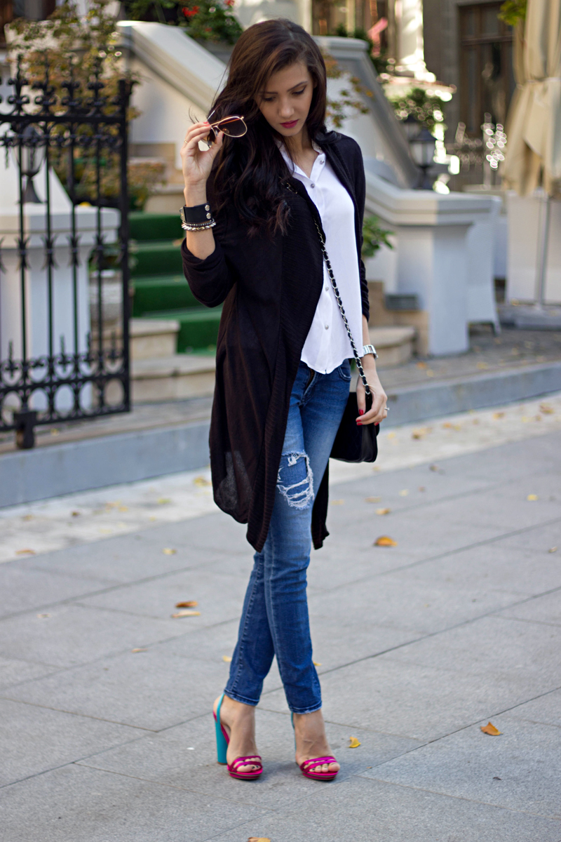 Woman wearing a longline black open cardigan over a white shirt with ripped blue jeans and colorful block heels