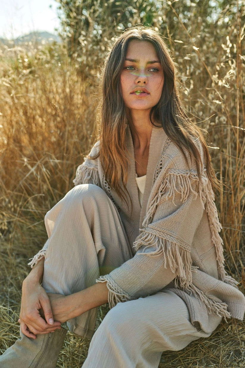 Woman sitting outdoors wearing a beige fringe-trimmed cardigan with matching beige wide-leg trousers