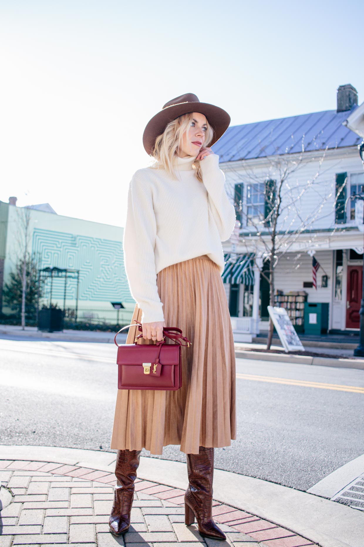 Woman wearing a cream turtleneck sweater with a camel pleated midi skirt, brown knee-high boots, a burgundy handbag and a brown fedora
