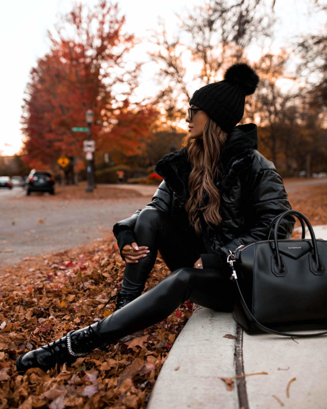Woman seated on a curb in a black puffer jacket, black leather leggings, black lace-up combat boots, beanie and sunglasses