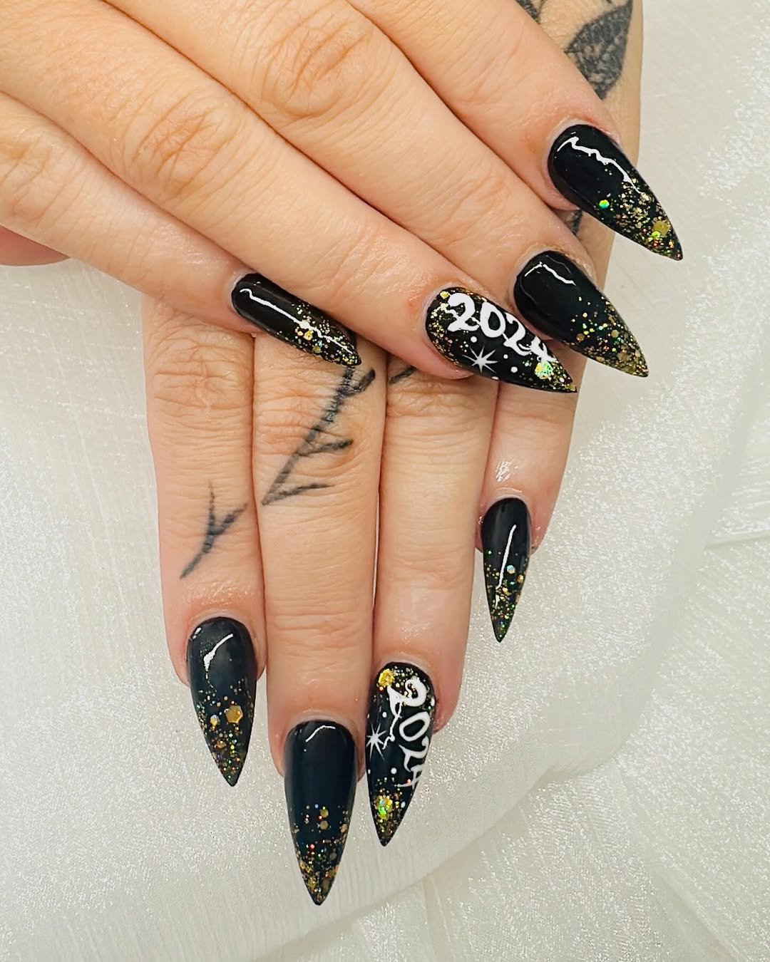 Black stiletto nails with gold glitter fading from the tips and white year-script art on accent nails