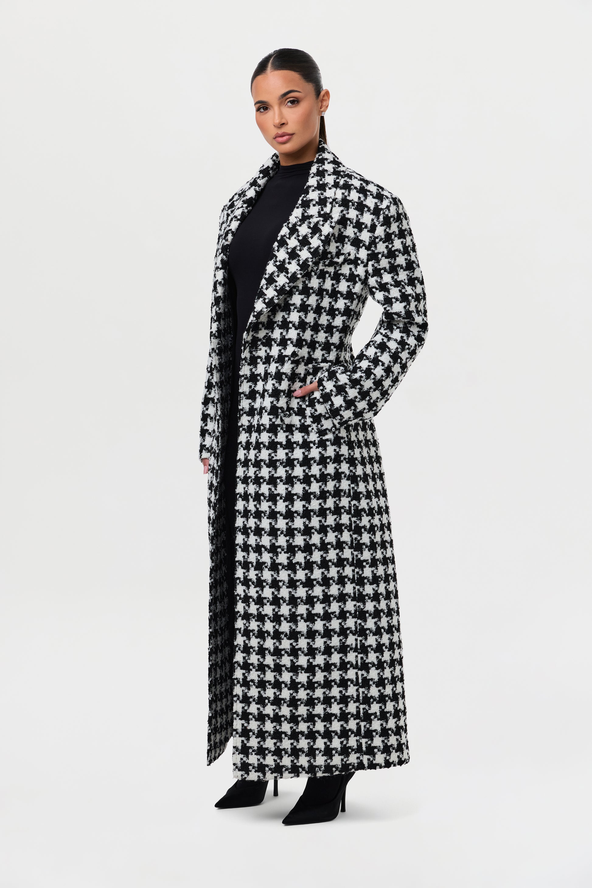 A black-and-white houndstooth long coat worn over black with pointed black stiletto boots.