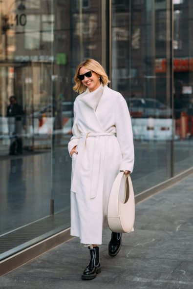 Woman in a white belted wrap coat with a white scarf, cream bag and black boots
