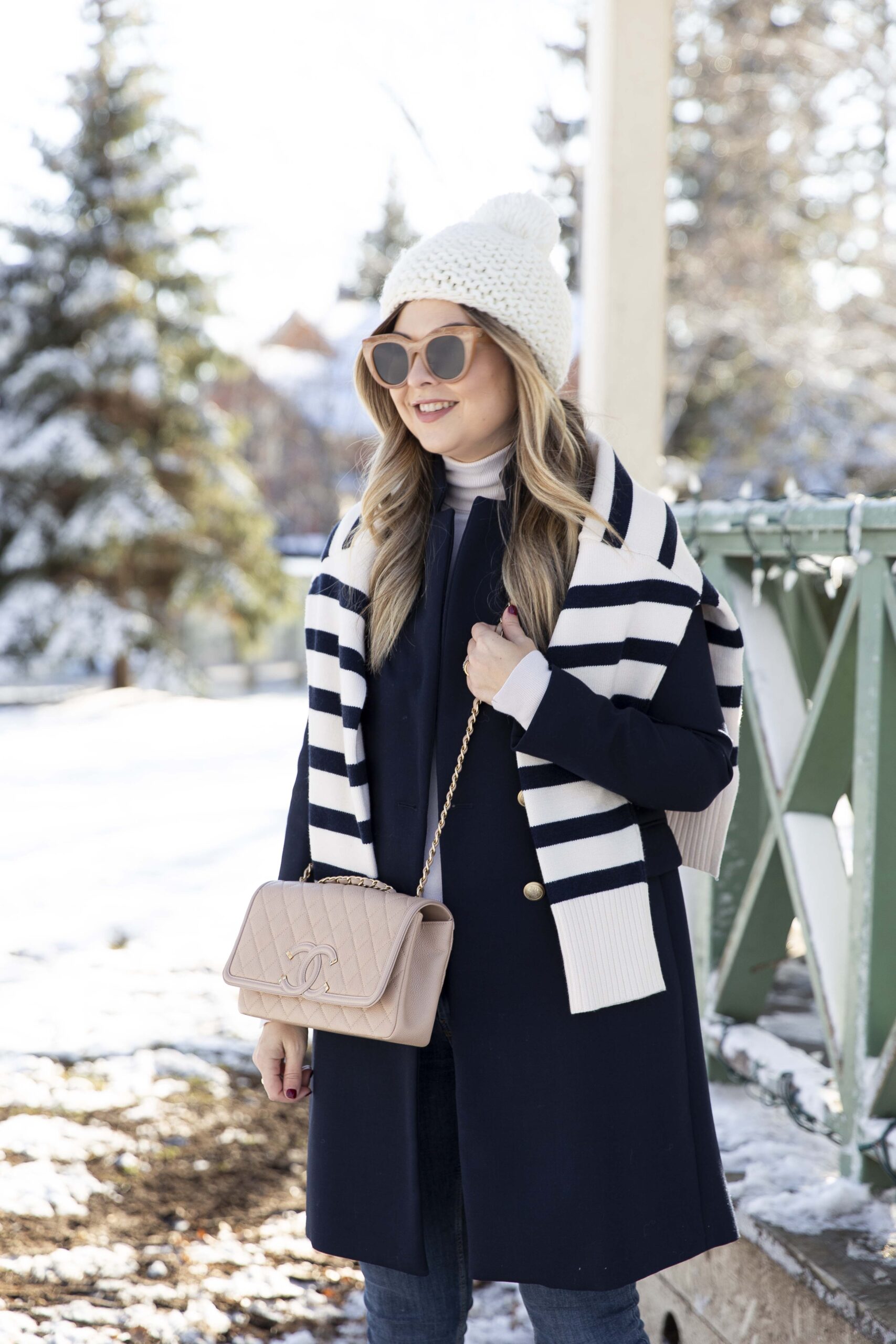 Woman with a navy and white striped scarf draped over a navy coat, white turtleneck and knit beanie in the snow