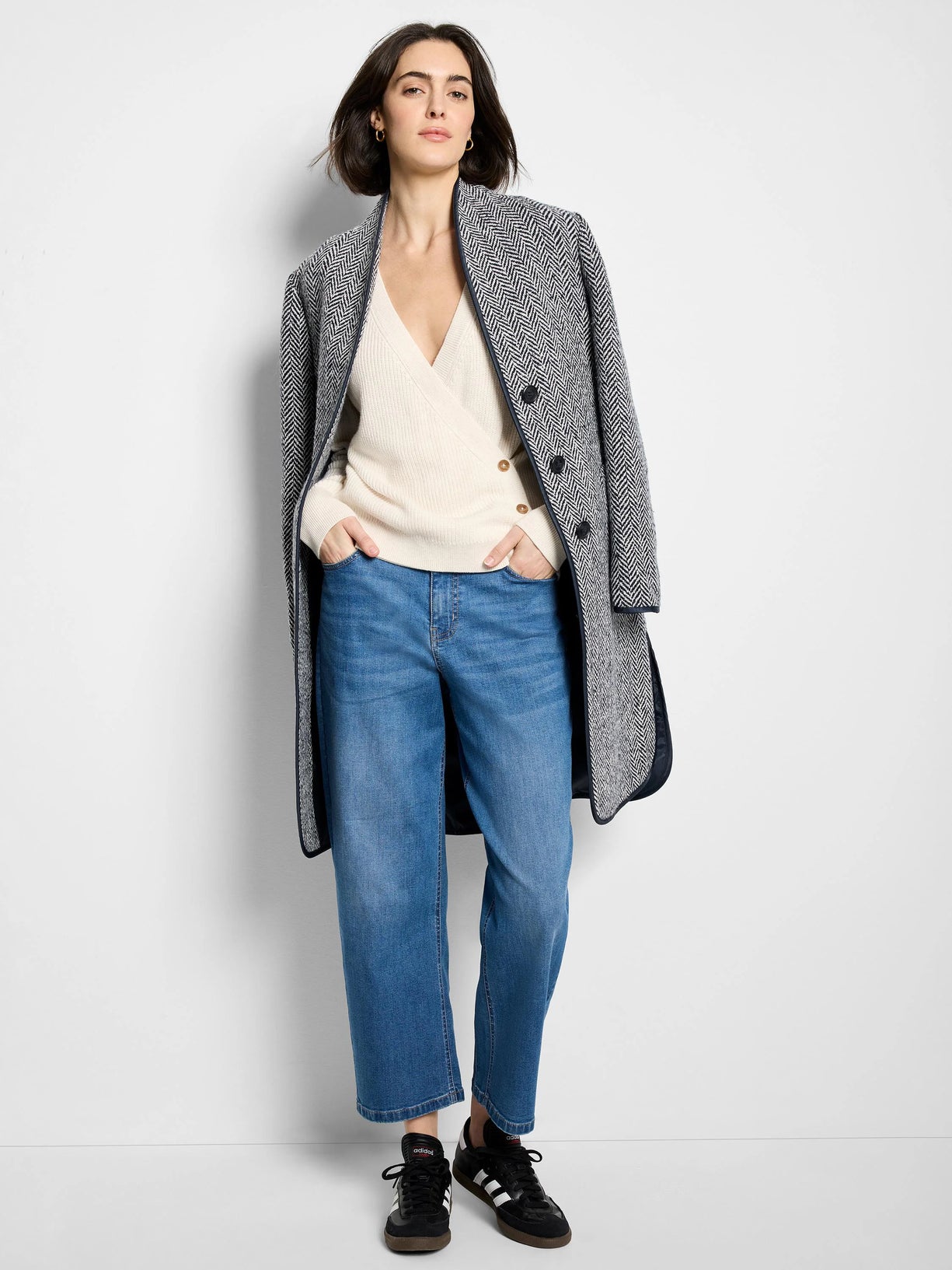 Woman in a grey herringbone wool top coat with cream wrap sweater, straight-leg jeans and black sneakers