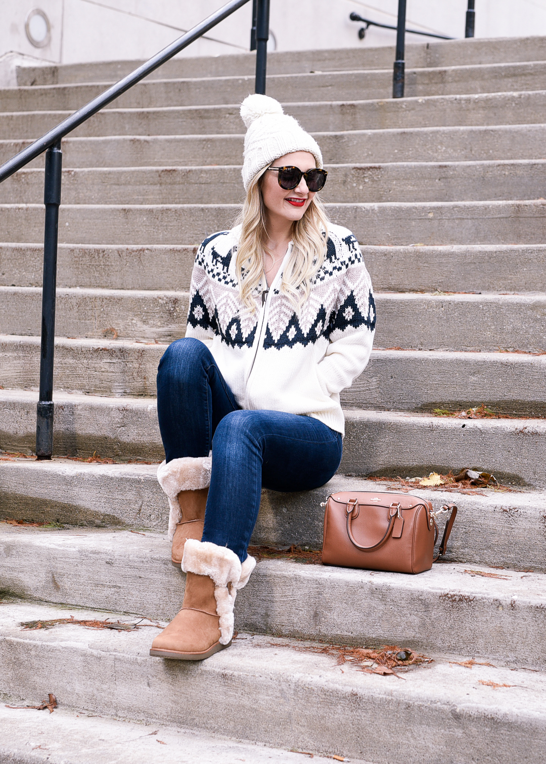 Woman in tan fur-trim boots, fair-isle sweater, dark jeans and a white pom beanie