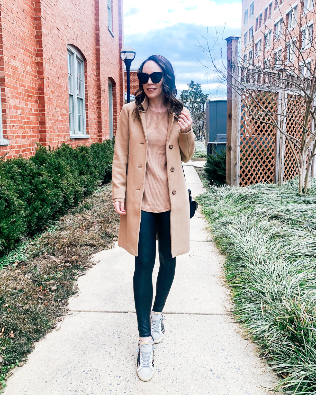 Woman in a camel coat, tan ribbed sweater, black leather leggings and sneakers