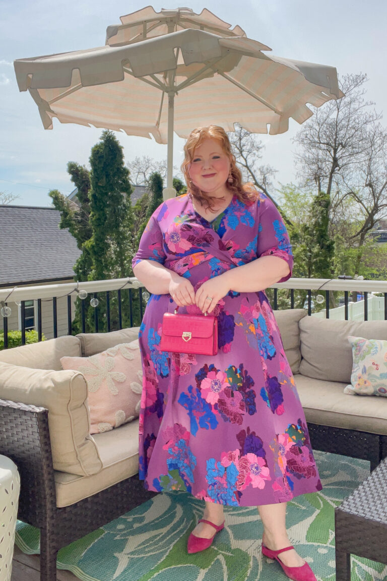Woman in a bold purple floral wrap midi dress with fuchsia bag and flats