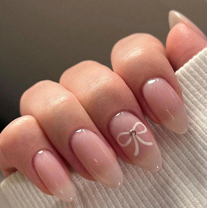 Short almond nude nails with a delicate white hand-painted bow and tiny gem center