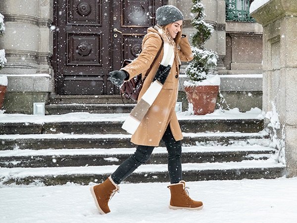 Woman in a camel coat and color-block scarf with brown suede winter boots in falling snow