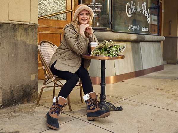 Woman sitting at a cafe table wearing a quilted jacket, leggings and two-tone lace-up snow boots