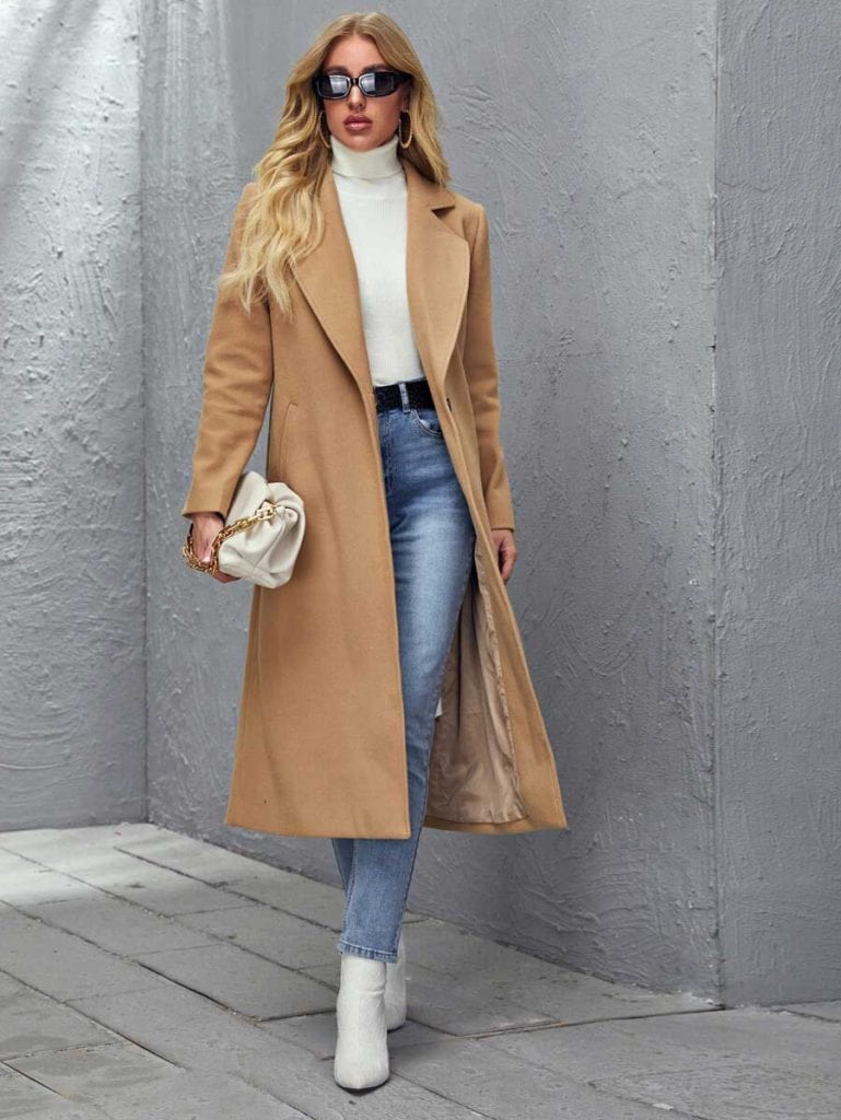 A camel long coat over a white turtleneck, blue jeans and white boots, with a cream pouch bag and sunglasses.