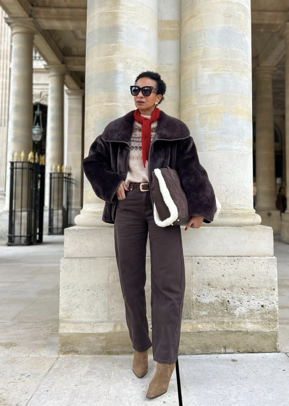 Dark shearling coat over fair-isle knit with red scarf, dark barrel-leg jeans and taupe suede boots