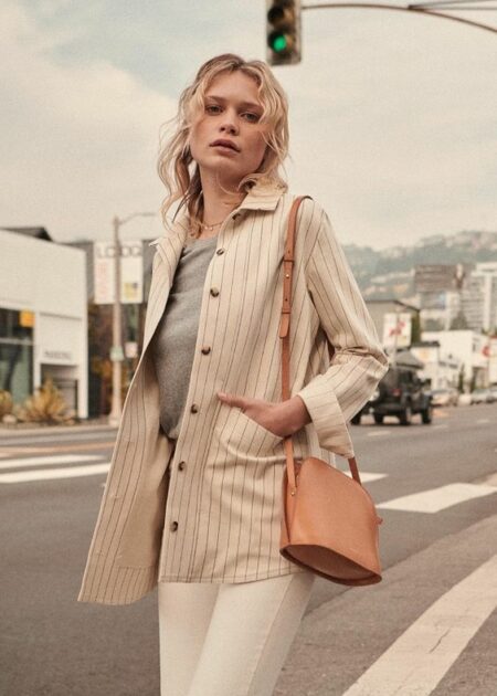 Woman wearing a cream pinstripe chore jacket over a gray top with white jeans and a tan shoulder bag