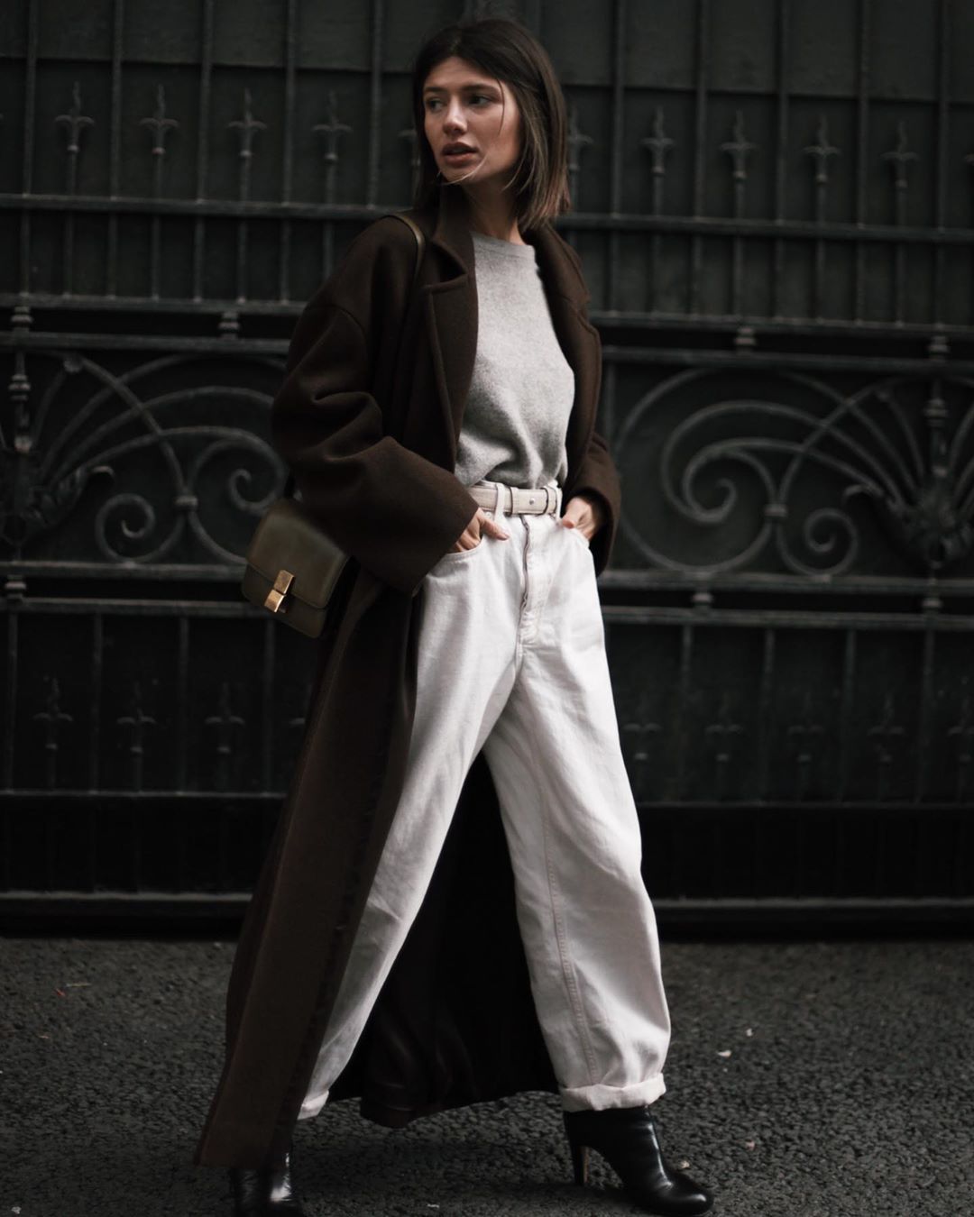 Woman wearing a dark brown maxi wool coat over a grey sweater and white jeans with black heeled ankle boots