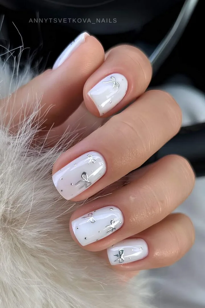 Short white nails with tiny silver bow charms on the ring fingers