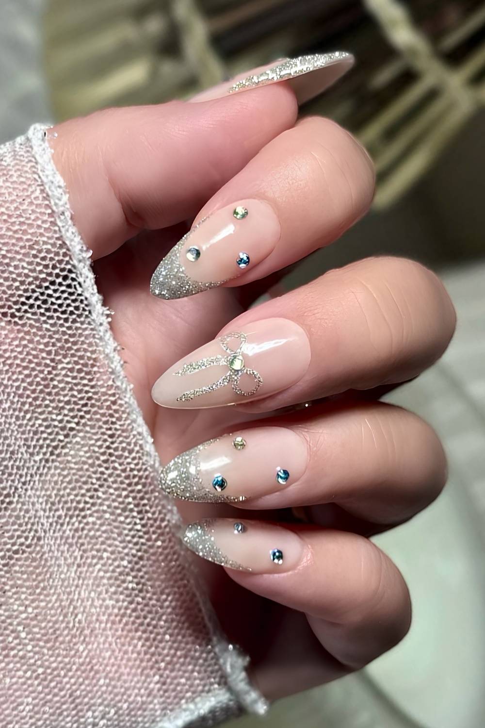 Almond nails with silver glitter French tips, a silver glitter bow design with a crystal center, and scattered blue gems