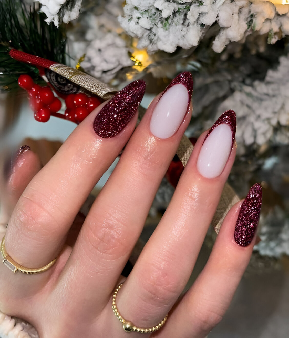 Almond nails with milky nude base, deep red glitter French tips and full red glitter accent nails against a Christmas tree